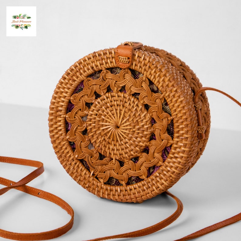 May include: A round, woven rattan bag with a brown leather strap. The bag features a decorative woven pattern on the front and a leather closure. The interior fabric is patterned with purple and gold designs. The bag is a natural brown color.