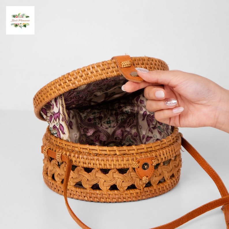 May include: A round, woven rattan bag with a brown leather strap and clasp. The bag's interior features a patterned fabric lining. The bag is open, revealing the interior. The bag is a natural tan color.