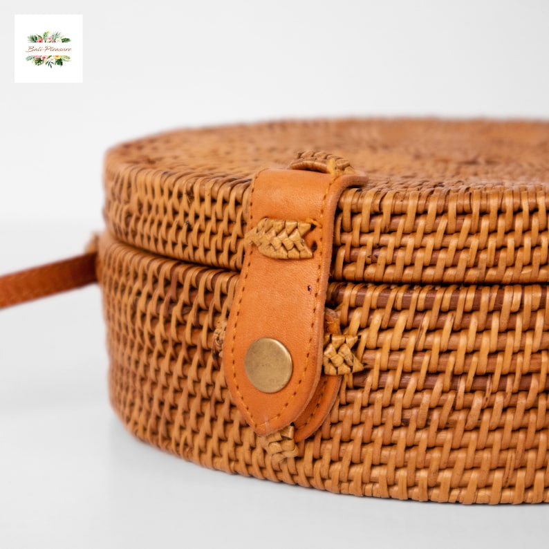 Op de afbeelding: Een ronde, geweven rotan handtas met een lichtbruine leren riem en sluiting. De tas heeft een geweven textuur en een messingkleurige knoopsluiting. De tas heeft een natuurlijke kleur en een eenvoudig, elegant ontwerp.