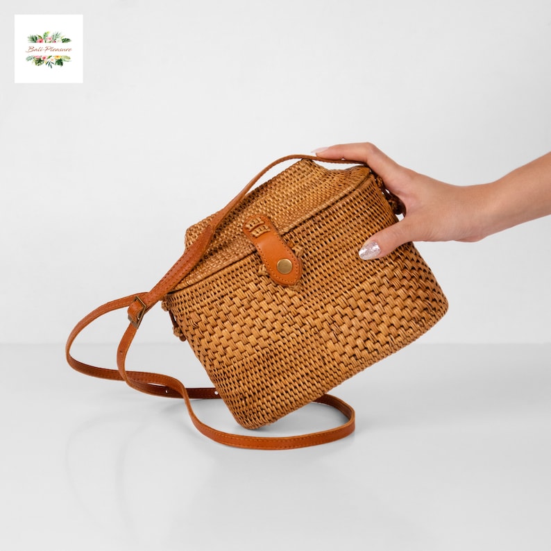 Peut inclure: Sac bandouli&egrave;re en rotin tress&eacute; de forme rectangulaire. Le sac est dot&eacute; d'une bandouli&egrave;re en cuir marron et d'une fermeture &agrave; rabat avec un bouton. Le sac est de couleur marron clair et a une surface textur&eacute;e. Le sac est tenu par une main.