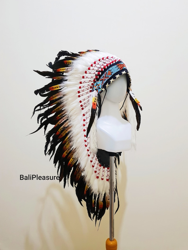 Op de afbeelding: Een Indiaanse hoofdtooi met witte, zwarte, gele en rode veren. De hoofdband is voorzien van kralen in blauw, rood en oranje. De tekst "BaliPleasure" is zichtbaar aan de linkerkant van de afbeelding.