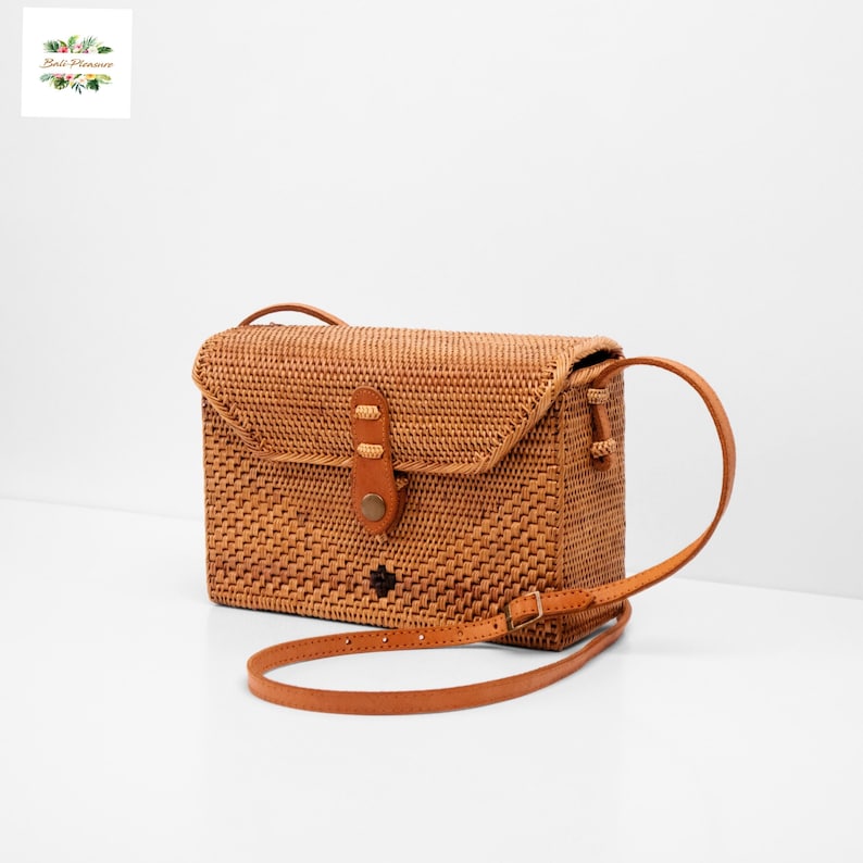 May include: A light brown rectangular crossbody bag made of woven rattan. The bag features a flap closure with a leather strap and buckle.  It has a short, adjustable leather shoulder strap. The bag is displayed on a white background.