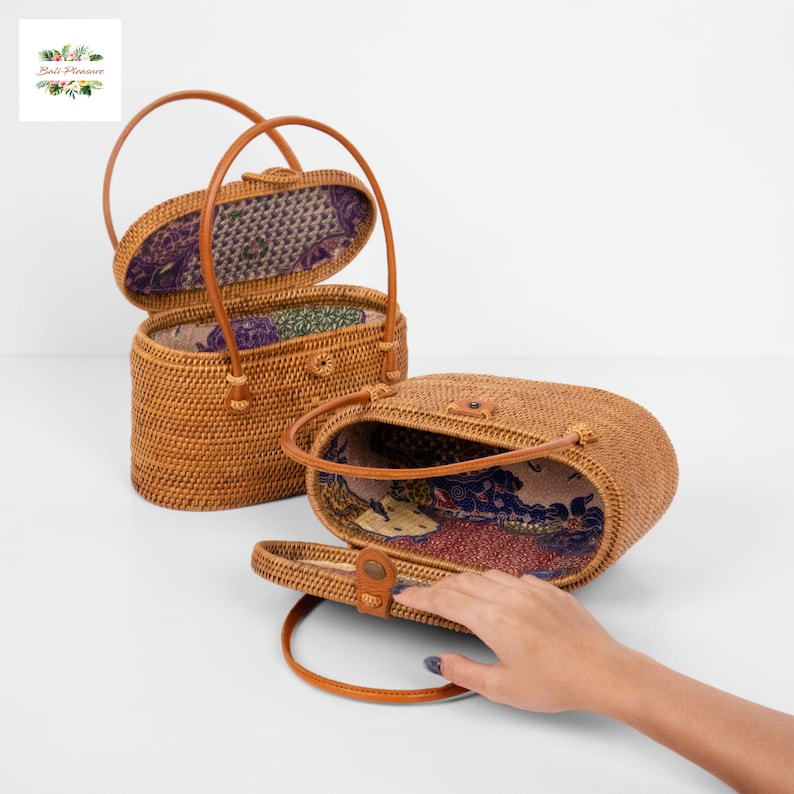 Puede incluir: Dos bolsos de mano ovalados de rat&aacute;n tejido con asas de cuero. Uno est&aacute; abierto, revelando un forro de tela estampada. El otro est&aacute; parcialmente abierto, mostrando el interior. Los bolsos son de color marr&oacute;n natural.