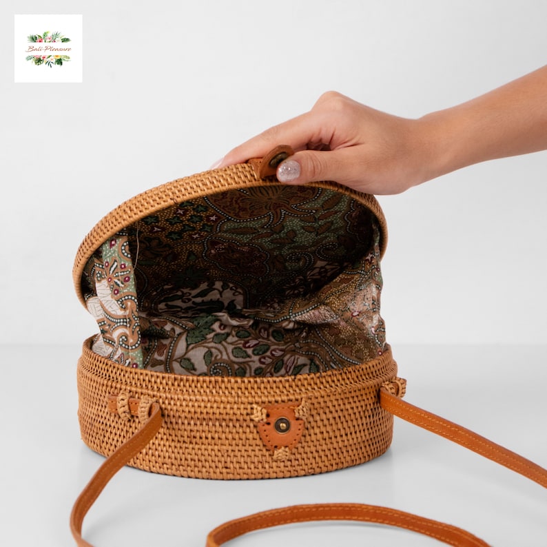 May include: A round, woven rattan handbag with a brown leather strap. The bag's interior features a patterned fabric lining in shades of green and brown. The bag is open, revealing the interior. The bag has a leather closure.