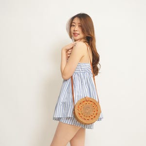 May include: A woman wearing a blue and white striped sundress and carrying a round, woven rattan crossbody bag with a brown leather strap. The bag has an intricate circular design.