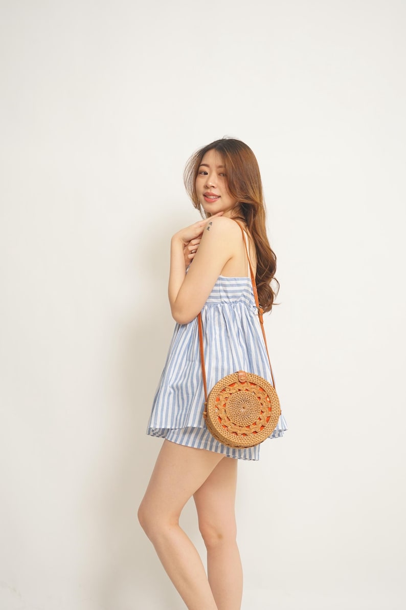 May include: A woman wearing a blue and white striped sundress and carrying a round, woven rattan crossbody bag with a brown leather strap. The bag has an intricate circular design.