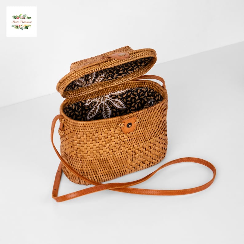 Peut inclure: Sac bandouli&egrave;re en rotin tress&eacute; de forme rectangulaire avec un couvercle &agrave; charni&egrave;re. Le sac est dot&eacute; d'une bandouli&egrave;re en cuir marron et d'une doublure en tissu &agrave; motifs. Le sac est ouvert, r&eacute;v&eacute;lant l'int&eacute;rieur. Le sac est de couleur beige naturel.