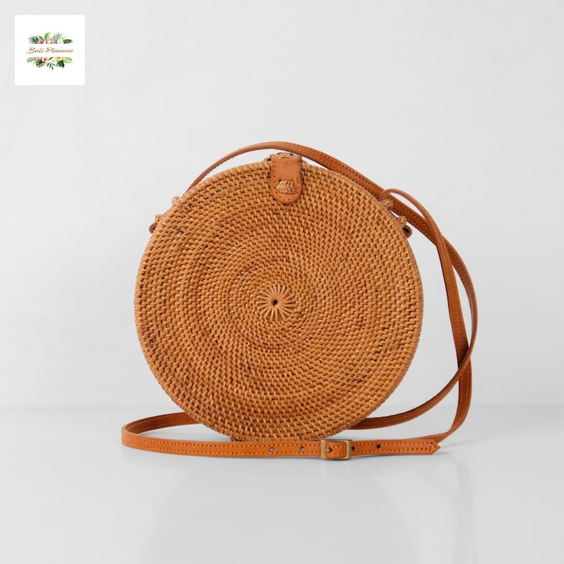 May include: A round, woven rattan crossbody bag with a brown leather strap and buckle. The bag has a spiral design in the center and a leather flap closure. The bag is a light brown color.
