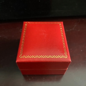 May include: A small, rectangular red box with a gold-colored, wavy border. The box is closed and appears to be made of a textured material. The box is sitting on a dark surface.