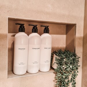 Op de afbeelding: Drie witte flessen met zwarte pompen, met de labels 'Shampoo', 'Conditioner' en 'Body Wash', staan in een nis. Elke fles heeft een minimalistisch etiket. Er is ook een kleine potplant met hangende groene bladeren.