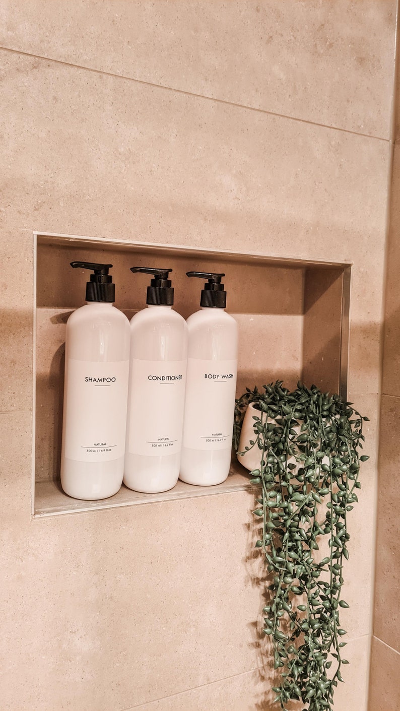 Op de afbeelding: Drie witte flessen met zwarte pompen, met de labels 'Shampoo', 'Conditioner' en 'Body Wash', staan in een nis. Elke fles heeft een minimalistisch etiket. Er is ook een kleine potplant met hangende groene bladeren.