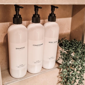 Op de afbeelding: Drie witte plastic flessen voor shampoo, conditioner en douchegel met zwarte pompen. Elke fles heeft een etiket met de productnaam. De flessen staan in een ingebouwde doucheruimte met een kleine plant in een pot.