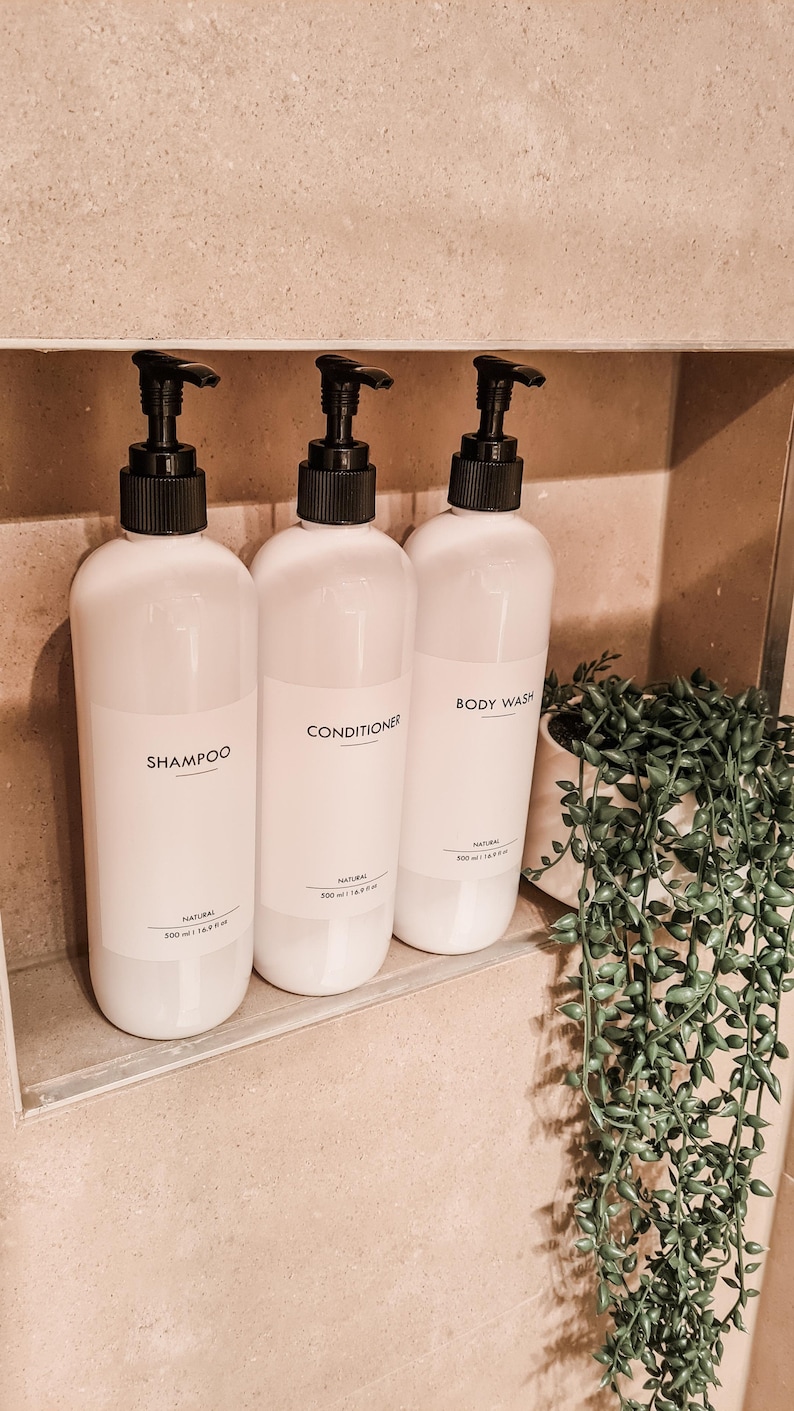 Op de afbeelding: Drie witte plastic flessen voor shampoo, conditioner en douchegel met zwarte pompen. Elke fles heeft een etiket met de productnaam. De flessen staan in een ingebouwde doucheruimte met een kleine plant in een pot.