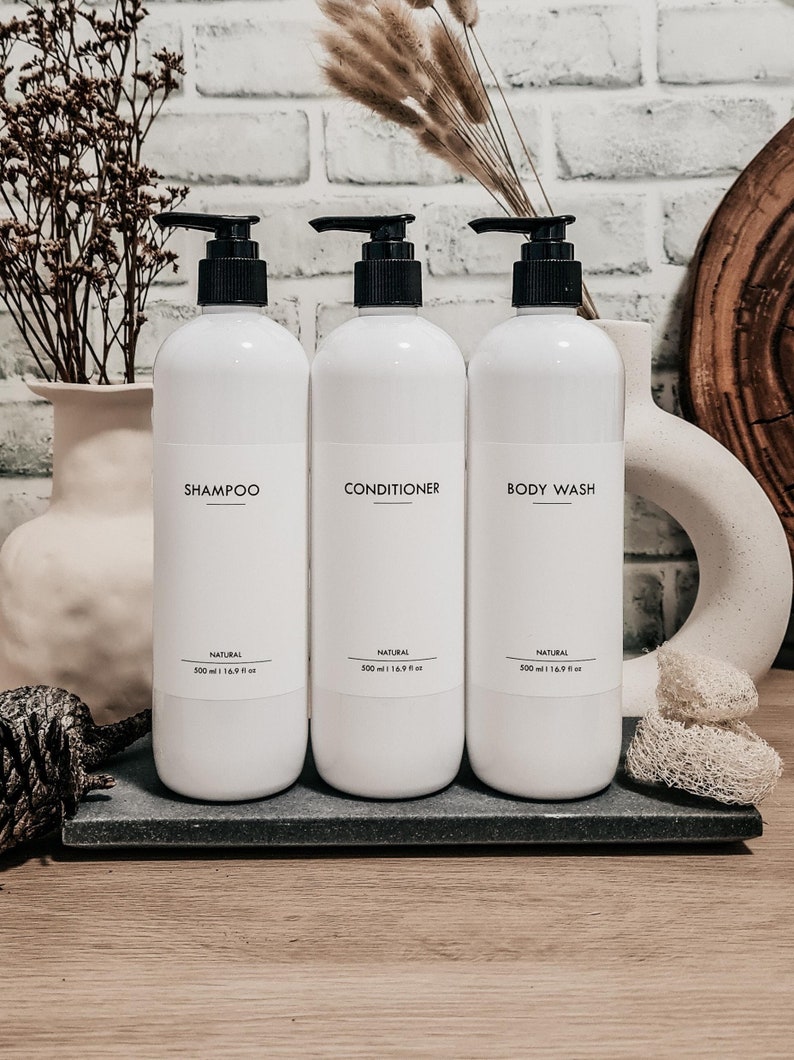 Op de afbeelding: Drie witte shampoo-, conditioner- en douchegelflessen met zwarte pompen. Elke fles heeft een etiket met de productnaam en de tekst "NATURAL". De flessen staan op een donkergrijze tray, met decoratieve items op de achtergrond.