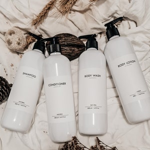 Op de afbeelding: Vier witte plastic flessen met zwarte pompkappen, met de labels "Shampoo", "Conditioner", "Body Wash" en "Body Lotion". Elke fles heeft een inhoud van 500 ml. De flessen zijn geplaatst op een wit, getextureerd oppervlak.