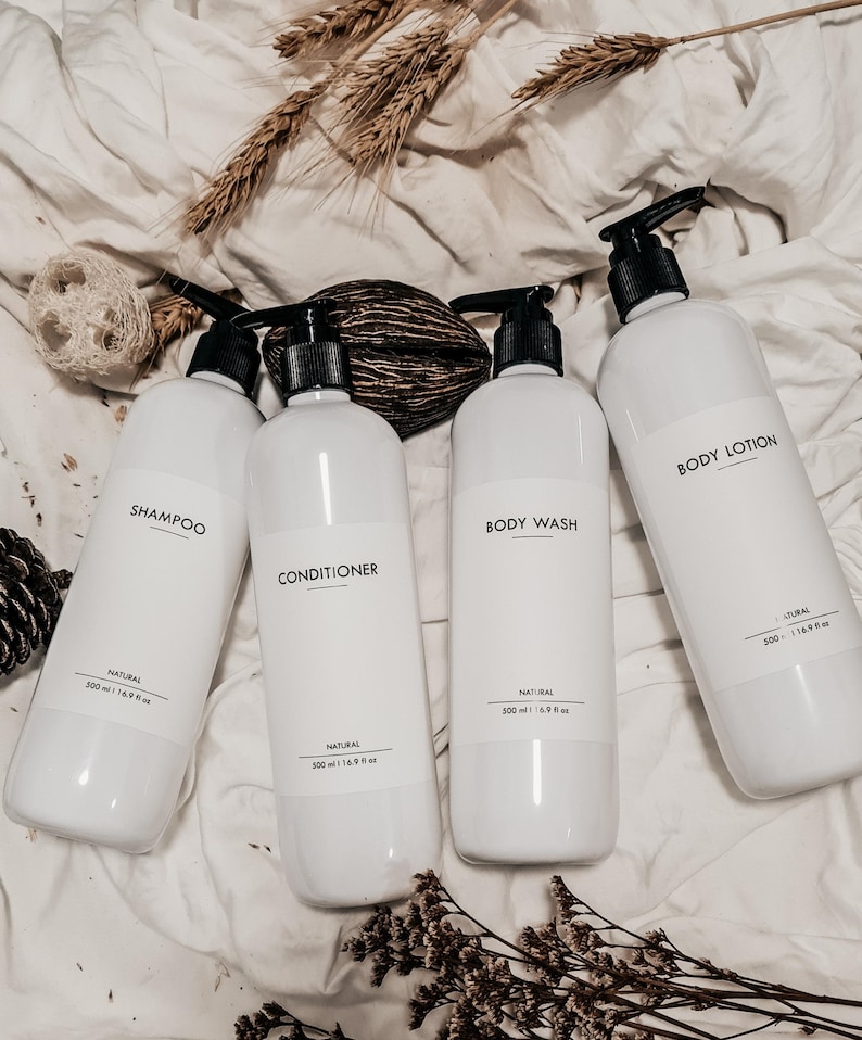 Op de afbeelding: Vier witte plastic flessen met zwarte pompkappen, met de labels "Shampoo", "Conditioner", "Body Wash" en "Body Lotion". Elke fles heeft een inhoud van 500 ml. De flessen zijn geplaatst op een wit, getextureerd oppervlak.