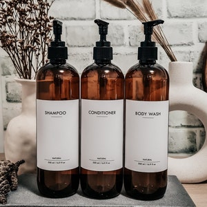 Op de afbeelding: Drie amberkleurige pompflessen met de labels "Shampoo", "Conditioner" en "Body Wash" op een grijze tray. Elke fles heeft een zwarte pomp en een wit label met zwarte tekst. De flessen zijn 500 ml.