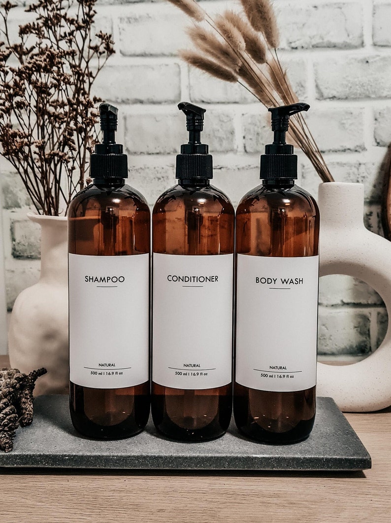 Op de afbeelding: Drie amberkleurige pompflessen met de labels "Shampoo", "Conditioner" en "Body Wash" op een grijze tray. Elke fles heeft een zwarte pomp en een wit label met zwarte tekst. De flessen zijn 500 ml.
