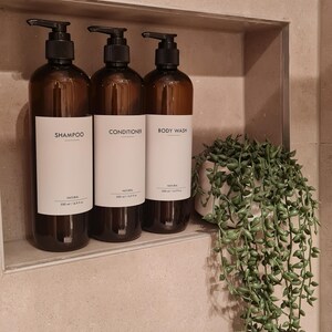 Op de afbeelding: Drie bruine amberglazen flessen met zwarte pompdispensers met de labels "SHAMPOO", "CONDITIONER" en "BODY WASH". Elke fles bevat 500 ml product. Rechts staat een kleine potplant met groene, trapsgewijze bladeren.
