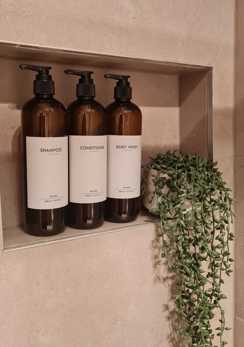 Op de afbeelding: Drie bruine amberglazen flessen met zwarte pompdispensers met de labels "SHAMPOO", "CONDITIONER" en "BODY WASH". Elke fles bevat 500 ml product. Rechts staat een kleine potplant met groene, trapsgewijze bladeren.