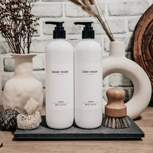 Op de afbeelding: Twee witte pompflessen met de opschriften "HAND WASH" en "DISH WASH" met zwarte pompen, elk met 500 ml inhoud. Een borstel met houten handvat en decoratieve items staan op een donkergrijze tray. Achtergrond: witte bakstenen muur.
