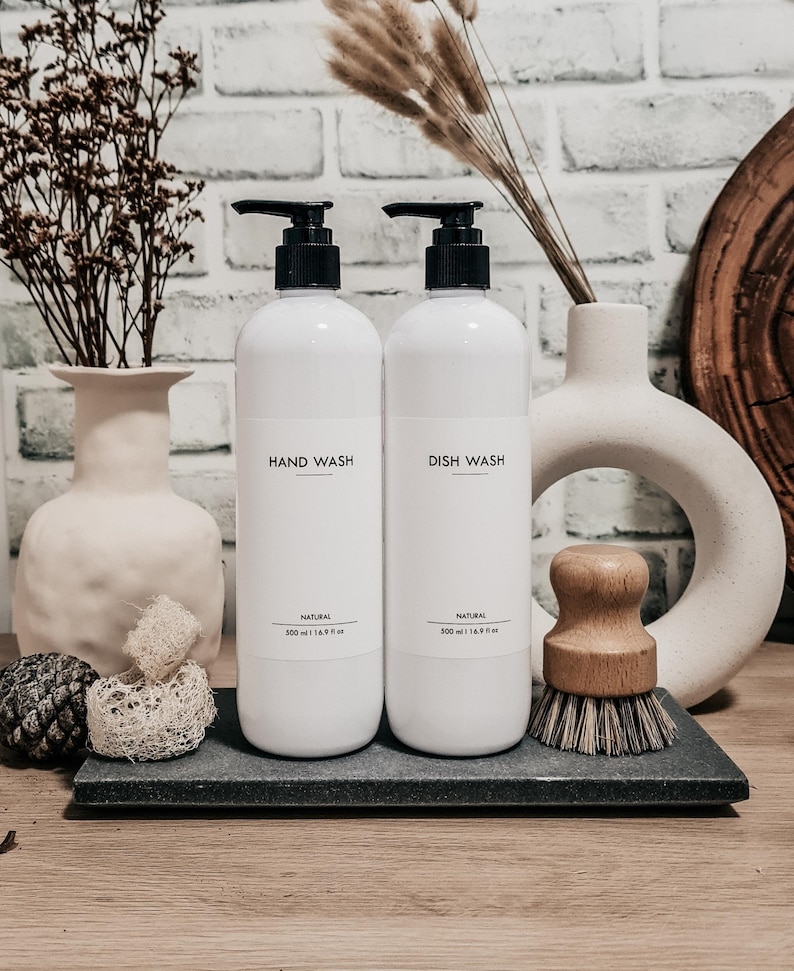 Op de afbeelding: Twee witte pompflessen met de opschriften "HAND WASH" en "DISH WASH" met zwarte pompen, elk met 500 ml inhoud. Een borstel met houten handvat en decoratieve items staan op een donkergrijze tray. Achtergrond: witte bakstenen muur.