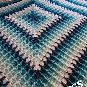 Puede incluir: Una manta de ganchillo hecha a mano en tonos de azul, turquesa y blanco. La manta presenta un patrón texturizado con un diseño de chevrón. La manta se muestra en una cama con almohadas. Las letras "TGS" están en la esquina inferior derecha.