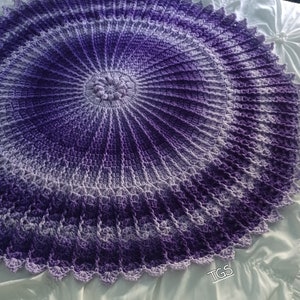 May include: A large, round, crocheted blanket in shades of purple and white. The blanket has a scalloped edge and a centre design that resembles a flower.