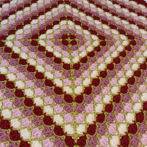 Puede incluir: Una manta de ganchillo hecha a mano con un patrón cuadrado. La manta presenta un patrón repetitivo de cuadrados en tonos burdeos, rosa, crema y verde. Los bordes son festoneados. Un artículo de decoración del hogar acogedor.