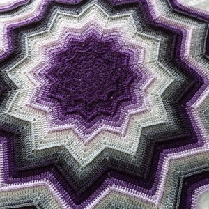 Puede incluir: Una manta de ganchillo hecha a mano con un diseño de estrella. La manta presenta un degradado de colores, comenzando con un centro morado oscuro y pasando por tonos de lavanda, gris y negro. La manta está hecha de hilo suave.