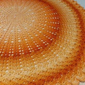 May include: A circular, crocheted blanket in shades of orange and yellow, creating a gradient effect. The handmade blanket features a detailed openwork pattern and a scalloped edge. The text "Twisted Granny at Home" is visible in the bottom right corner.