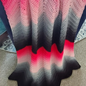 May include: A handmade crochet blanket with a chevron pattern. The blanket features a gradient of colours, starting with bright pink at the top, transitioning through lighter pinks, greys, and black at the bottom. The blanket has a scalloped edge.