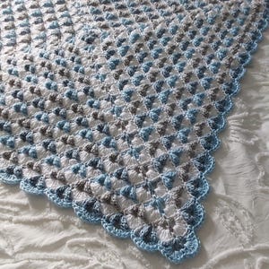 Könnte beinhalten: Eine quadratische Häkeldecke mit einem sich wiederholenden Granny-Square-Muster aus weißem, hellblauem und grauem Garn. Die Decke ist mit einem hellblauen, wellenförmigen Rand versehen. Der Text "Twisted Granny" steht unten im Bild.