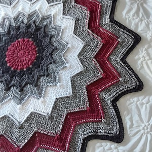 May include: A handmade crochet blanket with a starburst design. The blanket features a central red flower motif, surrounded by concentric rings of white, gray, and dark gray. The outer edge has a zigzag pattern in red, gray, and black.