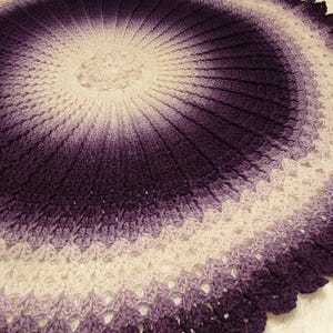 Puede incluir: Una manta redonda de ganchillo en tonos morados y blancos. El diseño irradia desde el centro, con un borde festoneado. El color transiciona de blanco en el centro a morado oscuro en el borde. Aproximadamente 100 cm de diámetro.