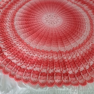 May include: A handmade, circular crochet blanket in shades of pink and coral. The blanket features a scalloped edge and a central floral design. The color gradient creates a soft, textured appearance. This item is perfect for home decor.