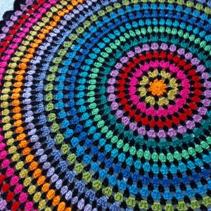 Puede incluir: Una manta circular de ganchillo con un patrón de arcoíris. La manta presenta anillos concéntricos de colores brillantes, incluyendo rojo, naranja, amarillo, verde, azul y morado, sobre un fondo negro. El texto "Twisted Granny" es visible.
