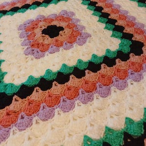 Può includere: Una coperta all'uncinetto con un motivo geometrico colorato. La coperta è realizzata con filato bianco, nero, verde, rosa e viola. La coperta ha un bordo a festoni.
