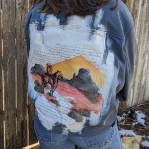 May include: A denim jacket with a hand-painted scene of a cowboy riding a horse through a desert landscape. The jacket has a white background with a blue denim overlay. The text "Janeury, baby, the part of me apart from me" is written on the jacket.