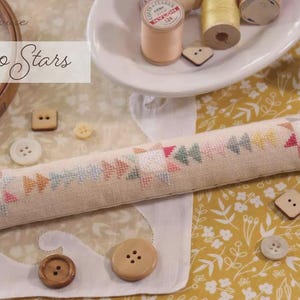 May include: A long, rectangular, beige fabric pincushion featuring a cross-stitched pattern of stars and geometric shapes in pink, yellow, green, and blue. The text October House Fiber Arts Calico Stars OH1300 is visible in the upper left corner.