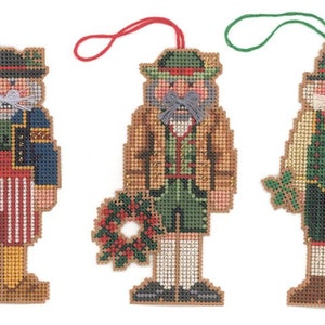 May include: Three hand-stitched felt ornaments depicting traditional German Christmas figures. Each ornament features a different figure wearing a festive costume and holding a unique accessory. The figures are dressed in red, green, and blue with white accents.