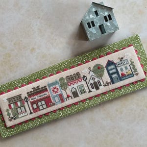 May include: A cross-stitch design depicting a quaint town scene with buildings, trees, and streetlights. The design is stitched on a beige fabric and framed with a red scalloped border. The background is a green fabric with a white pattern. A small metal house is in the background.