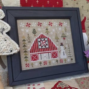 May include: Framed cross-stitch artwork featuring a red barn with a star design, a snowman, and Christmas trees. The artwork is set against a beige background with snowflakes and is enclosed in a dark blue frame. The overall theme is a winter holiday scene.