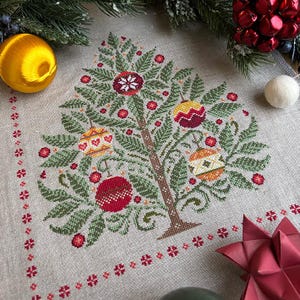 May include: A cross-stitch design of a Christmas tree with ornaments. The tree is brown with green leaves and red, yellow, and white ornaments. The design is on a beige fabric with a red floral border. A gold ornament and greenery are in the background.