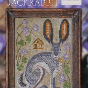 May include: Cross-stitch sampler titled "The Jackrabbit" from "A Year in the Woods #3." The framed artwork features a jackrabbit in shades of gray, orange, and white, surrounded by purple flowers and a small house. The frame is a dark brown.