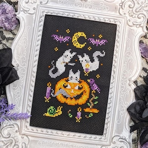May include: Cross-stitch art in a white ornate frame. The black fabric features a Halloween design with a smiling jack-o'-lantern, two ghost cats, bats, and a crescent moon. The artwork includes purple, orange, yellow, and white details, creating a festive seasonal theme.