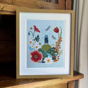 May include: Framed cross-stitch artwork featuring a windmill, flowers, and a small house. The artwork is set against a light blue background and framed in a light wood frame. The flowers include red poppies, white daisies, and orange blooms.