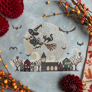 May include: Cross-stitch design featuring a witch on a broom with a black cat, flying in front of a full moon. Below, a row of houses and trees are stitched in detail. The color palette includes shades of blue, green, and brown.
