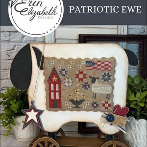 May include: A wooden sheep-shaped decorative piece with a stitched design featuring a red house, stars, and an American flag. The piece is mounted on a wooden cart with wheels and includes a star-shaped ornament. The text "PATRIOTIC EWE" is displayed.