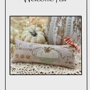 May include: A rectangular, cross-stitched pillow with the words "Welcome Fall" and a pumpkin design. The pillow is beige with brown and green stitching, and a small orange and white bow. Two pumpkins are in the background.